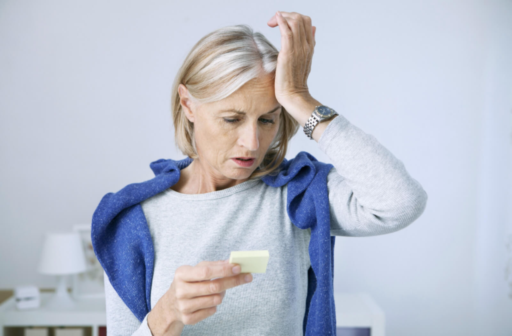 A senior woman in a sweater puts the heel of her palms on her forehead as she suddenly remembers something important after seeing a reminder on a small sticky note.