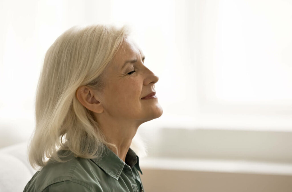 A close-up side view of a tranquil senior woman with blonde hair meditating at home, sitting peacefully with her eyes closed.