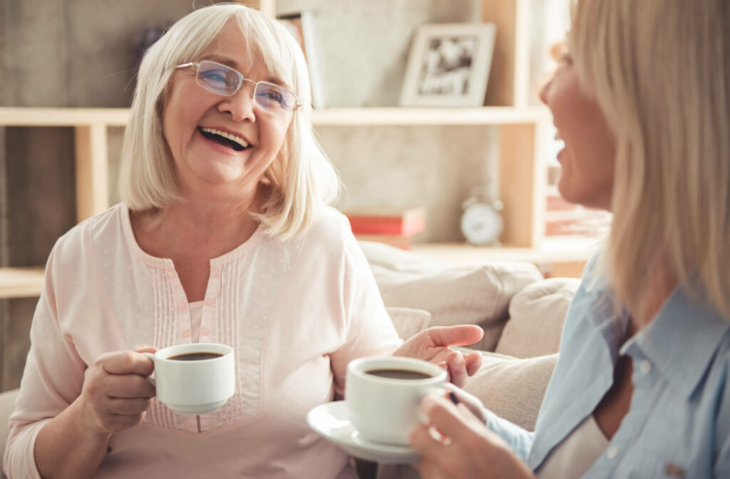 An older adult woman and her daughter sitting on a couch smiling and talking to each other while holding a cup of tea.