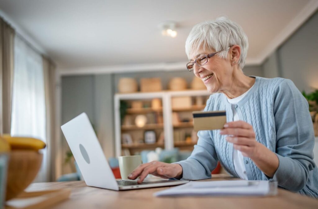 An older adult woman holding a credit or debit card on her left hand while doing online shopping using a laptop computer