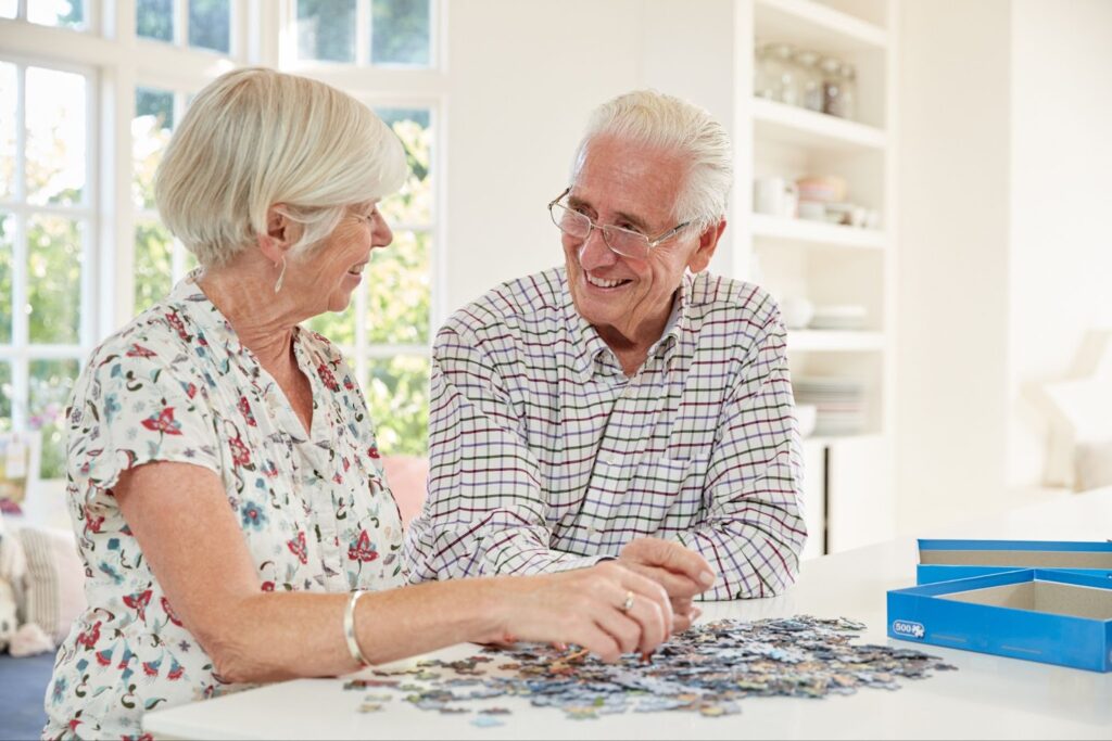 A senior couple smiling at each other while doing a puzzle together.