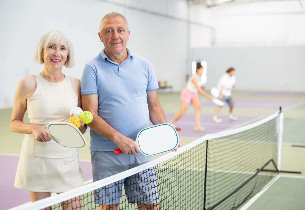 A smiling pair of seniors holding pickleball equipment on an indoor court.
