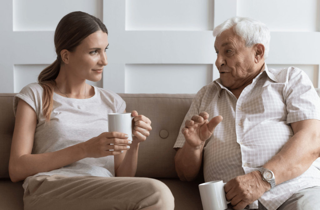 n adult child sits with their senior parent, discussing something over coffee.