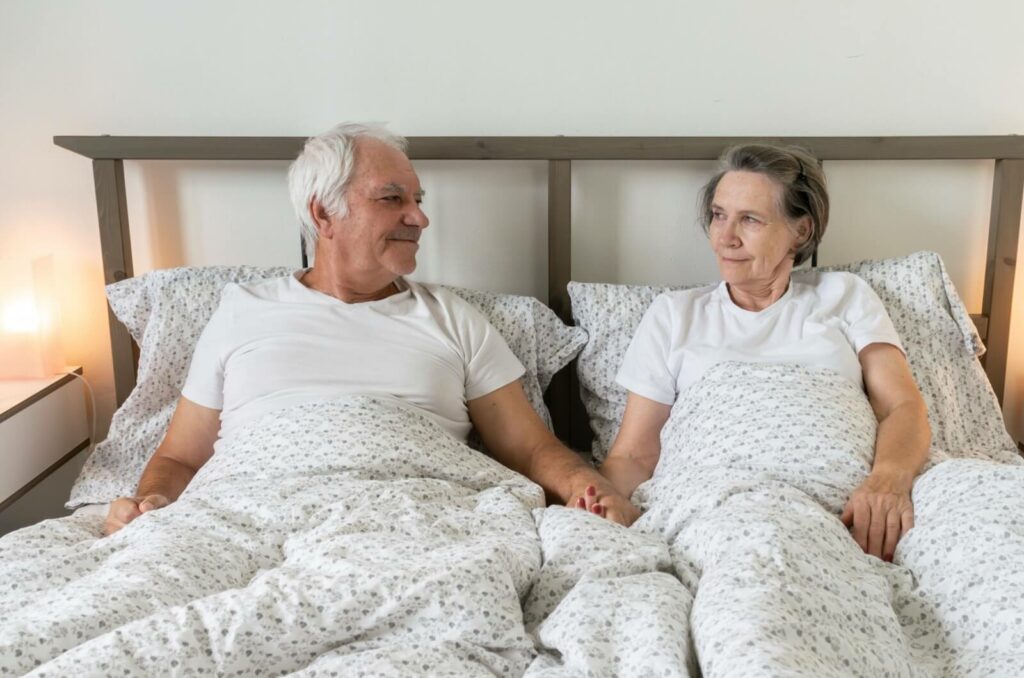 A smiling older couple in bed looking at each other and holding hands.